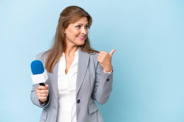 İzole edilmiş mavi arkaplan üzerinde orta yaşlı TV sunucusu kadın bir ürünü sunmak için kenarı işaret ediyor