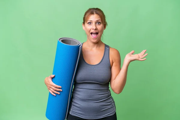 Middle-aged sport woman going to yoga classes while holding a mat over isolated background with shocked facial expression