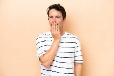 Beyazlı genç bir adam bej arka planda izole edilmiş. Ağzını eliyle kapatıyor.