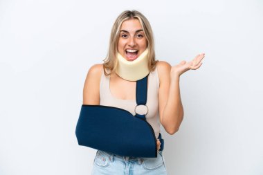 Kırık kollu ve askı takan bir kadın beyaz arka planda izole edilmiş, şok olmuş bir yüz ifadesi var.