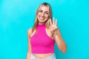 Mavi arka planda izole edilmiş genç beyaz kadın mutlu ve parmaklarıyla üçe kadar sayıyor.