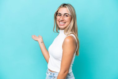 Mavi arka planda izole edilmiş genç beyaz kadın, davet edilmek için ellerini yana uzatıyor.