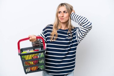 Young caucasian woman holding a shopping basket full of food isolated on white background having doubts