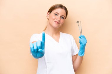 Beyaz dişçi kadın, elinde bej arka planda izole edilmiş aletler tutuyor ve parmağını kaldırıyor.
