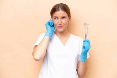 Elinde bej arka planda izole edilmiş aletler tutan beyaz dişçi kadın hayal kırıklığına uğramış ve kulakları tıkamış.