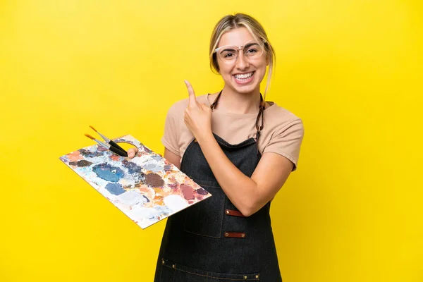黄色の背景に孤立したパレットを持っている若い芸術家の白人女性は 製品を提示する側を指している — ストック写真