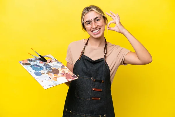 黄色の背景に孤立したパレットを持っている若いアーティストCaucasian女性は指でOkのサインを示す — ストック写真