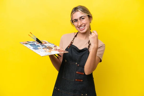 黄色の背景に孤立したパレットを保持する若いアーティストの白人女性がやって来るジェスチャーを行う — ストック写真
