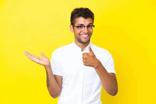 若いですハンサムなブラジル人男性孤立した黄色の背景に保持コピースペース想像上のヤシの上に挿入する広告と親指アップ — ストック写真