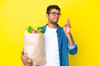 Sarı arka planda izole edilmiş alışveriş torbası tutan Brezilyalı genç adam şans diliyor.