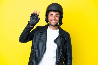 Motosiklet kasklı genç Brezilyalı adam sarı arka planda izole edilmiş mutlu bir ifadeyle selamlıyor.