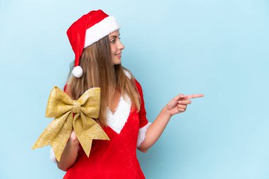 Anne Noel gibi giyinmiş genç beyaz kadın elinde Noel yayıyla mavi arka planda izole edilmiş bir şekilde bir ürünü sunmak için kenarı işaret ediyor.