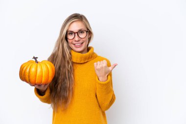 Beyaz arka planda izole edilmiş bir balkabağı tutan genç sarışın kadın bir ürünü sunmak için kenarı işaret ediyor.