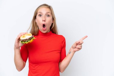 Beyaz arka planda izole edilmiş bir burger tutan genç sarışın kadın şaşırmış ve işaret ediyor.