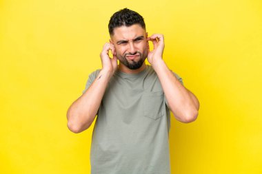Sarı arka planda izole edilmiş genç Arap yakışıklı adam hayal kırıklığına uğramış ve kulaklarını kapatmış.