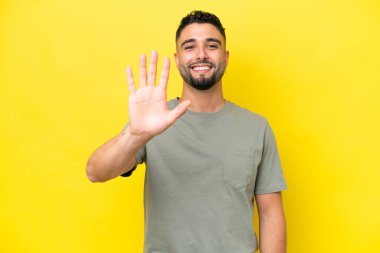 Sarı arka planda izole genç Arap yakışıklı adam parmaklarıyla beşi sayıyor.