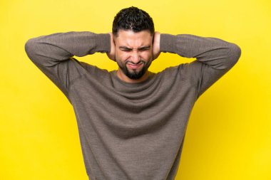 Sarı arka planda izole edilmiş genç Arap yakışıklı adam hayal kırıklığına uğramış ve kulaklarını kapatmış.