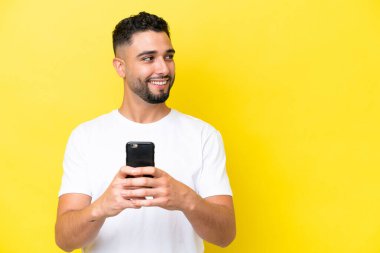 Young Arab handsome man isolated on yellow background using mobile phone and looking up