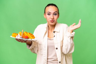 İzole edilmiş arka planda waffle tutan genç bir Arap kadını şüphe içinde el kaldırırken...