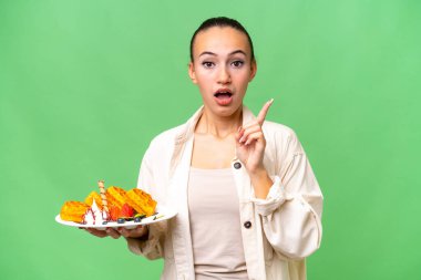 İzole edilmiş arka planda waffle tutan genç bir Arap kadını çözümü bulmak için parmağını kaldırıyor.