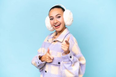 Mavi arka planda izole edilmiş, önü göstererek gülümseyen, kışlık kuklalar giyen genç bir Arap kadın.