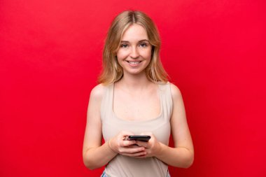 Kırmızı arka planda izole edilmiş genç bir İngiliz kadın cep telefonuyla mesaj gönderiyor.