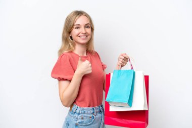 Beyaz arka planda izole edilmiş genç bir İngiliz kadın elinde alışveriş torbaları ve başparmağıyla