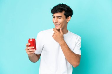 Arjantinli genç adam mavi arka planda izole edilmiş bir içecek tutuyor. Yan tarafa bakıyor ve gülümsüyor.