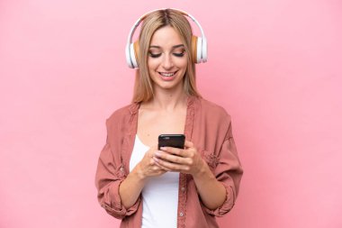 Güzel sarışın kadın pembe arka planda izole edilmiş müzik dinliyor ve cep telefonu arıyor.