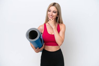 Genç sarışın kadın yoga derslerine gidiyor beyaz arka planda izole edilmiş bir minder tutarken yan tarafa bakıyor ve gülümsüyor.