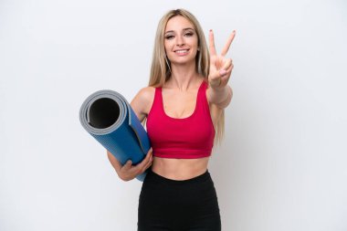 Genç sarışın spor kadın yoga derslerine gidiyor beyaz arka planda izole edilmiş bir minder tutarken gülümsüyor ve zafer işareti gösteriyor.