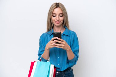 Güzel sarışın kadın beyaz arka planda izole edilmiş alışveriş torbaları tutuyor ve bir arkadaşına cep telefonuyla mesaj yazıyor.