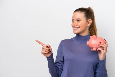 Beyaz arka planda izole edilmiş domuz kumbarası tutan genç beyaz bir kadın bir ürünü sunmak için kenarı işaret ediyor.