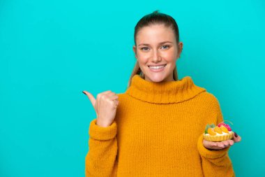 Mavi arka planda izole edilmiş turta tutan beyaz bir kadın bir ürünü sunmak için yan tarafı işaret ediyor.