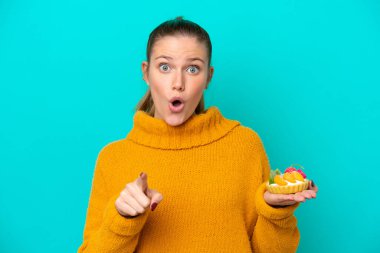 Mavi arka planda izole edilmiş tart tutan beyaz bir kadın şaşırmış ve ön tarafı işaret ediyor.