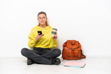 Beyaz arka planda bir katta oturan beyaz tenli genç bir kadın elinde kahve ve bir şeyler düşünürken bir yandan da cep telefonu tutuyor.