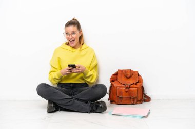 Beyaz arka planda beyaz bir zeminde oturan genç beyaz kadın şaşırdı ve bir mesaj gönderiyor.