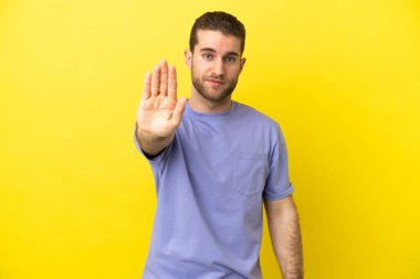 Sarışın yakışıklı adam, izole edilmiş sarı arka planda dur hareketi yapıyor.