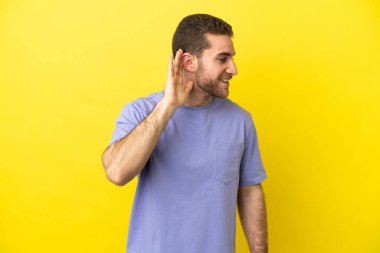 Handsome blonde man over isolated yellow background listening to something by putting hand on the ear