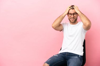 Genç adam pembe arka planda bir sandalyede oturmuş gülüyor.