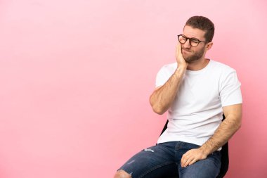 İzole edilmiş pembe arka planda diş ağrısı olan bir sandalyede oturan genç.