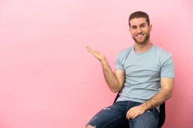 İzole edilmiş pembe arka planda bir sandalyede oturan genç adam bir ilan vermek için fotokopi makinesini avucunun içinde tutuyor.