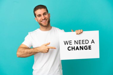 Handsome blonde man over isolated blue background holding a placard with text We Need a Change and  pointing it