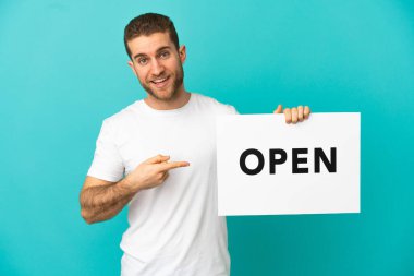 Yakışıklı, mavi arka planda, üzerinde açık yazı olan bir pankart tutan ve onu işaret eden bir sarışın.