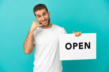 Yakışıklı sarışın, mavi arka planda. Elinde açık yazılı bir pankart var ve telefon hareketi yapıyor.