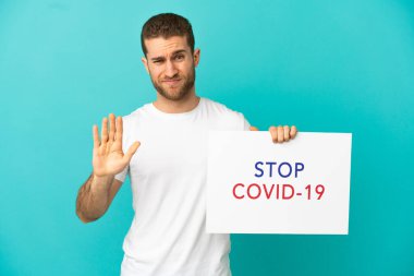 Yakışıklı sarışın, mavi arka planda. Elinde yazılı bir levha var. Covid 19 'u durdur ve dur işareti yap.