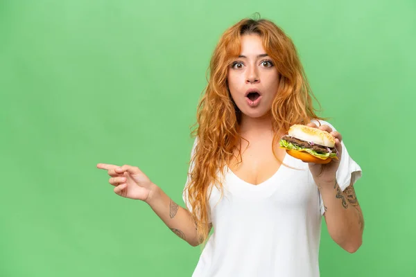 若いですCaucasian女性を保持していますハンバーガー隔離上の緑の画面クロマキー背景驚きと指摘側 — ストック写真