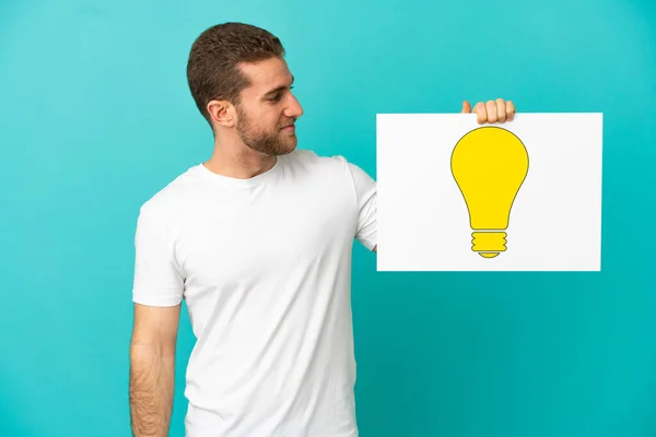 Handsome Blonde Man Isolated Blue Background Holding Placard Bulb Icon — Stock Photo, Image
