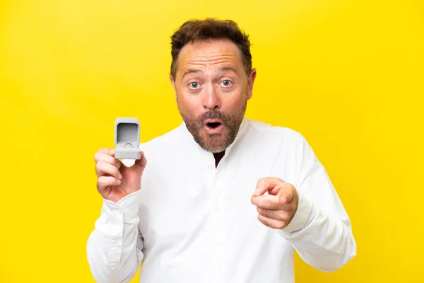 stock image Middle age man holding a engagement ring isolated on yellow background surprised and pointing front