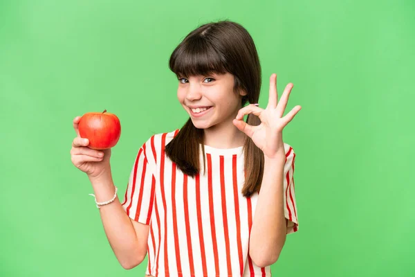 Little caucasian girl holding an apple over isolated background showing ok sign with fingers